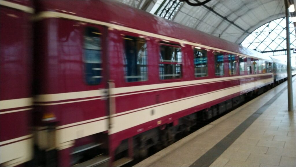 Dunkelroter Waggon mit weißen Streifen fährt am Bahnsteig ein. Rechts oberhalb sieht man Teile des weißen Daches vom Hauptbahnhof Dresden.