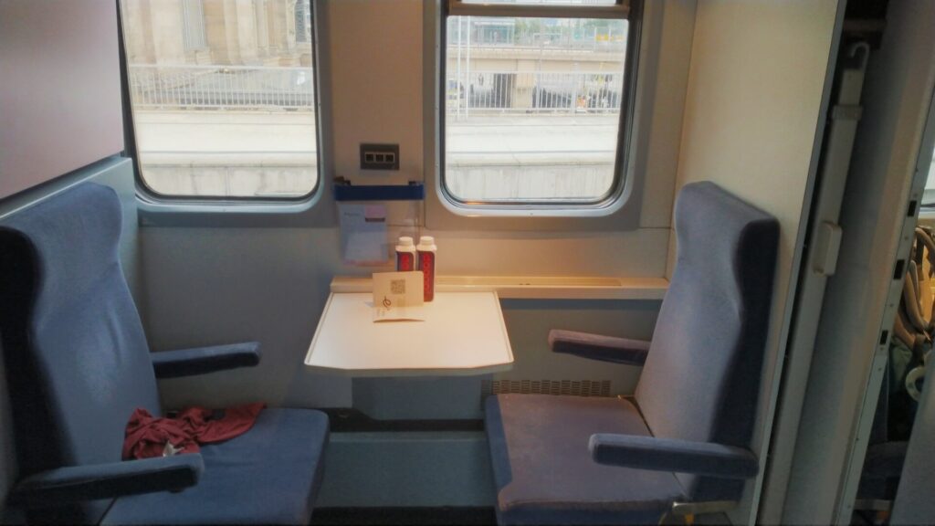 Zwei Sitze im European Sleeper Schlafwagen, dazwischen ein Tisch. Durch die zwei Fenster sieht man einen Bahnsteig.