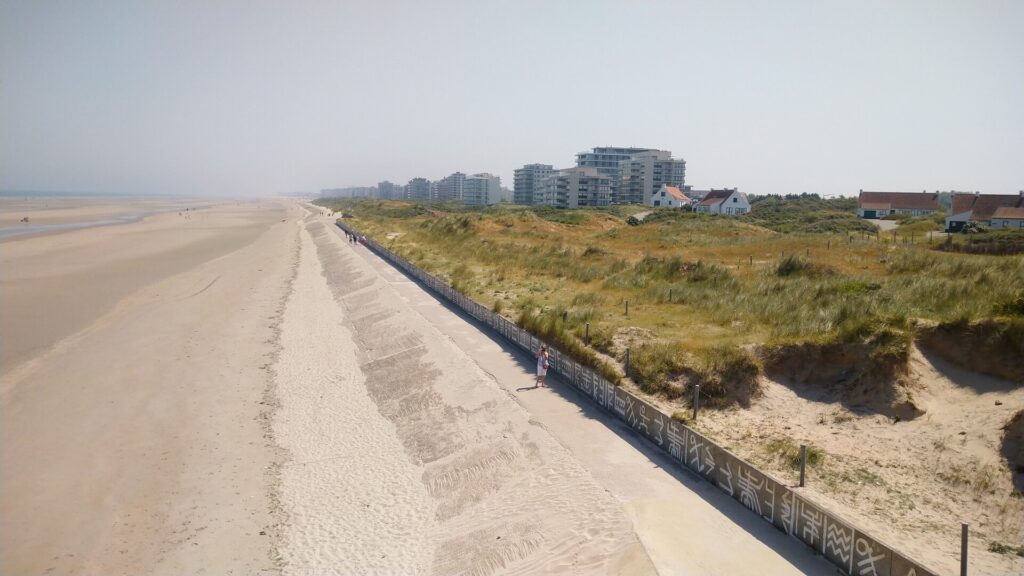 Links die Nordsee und ein breiter Strand, rechts hinten die Häuser, im Vordergrund rechts ein paar Dünen. Eine Mauer ist zwischen den Dünen und dem Strand.
